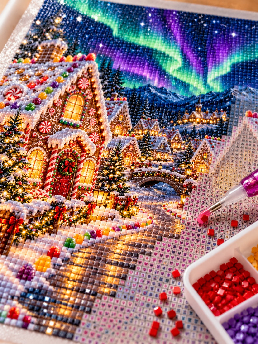 Close-up of the gingerbread village under green and violet northern lights diamond painting in progress, square resin drills, wax pen and drill tray
